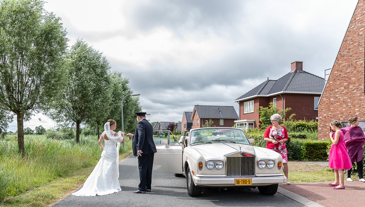 Bruidsfotografie; trouwfotograaf; De chauffeur van de trouwauto haalt de bruid op bij haar huis. De moeder en dochters van de bruid kijken toe.