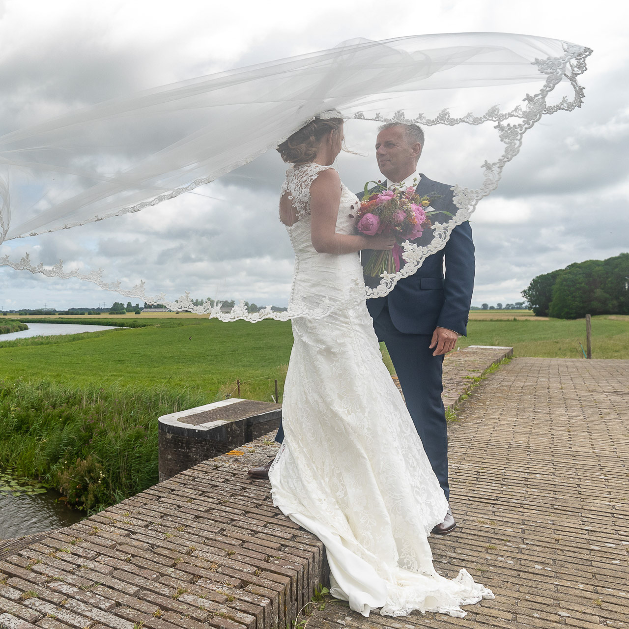 Bruidsfotografie; trouwfotograaf; De sluier van de bruid waait mooi omhoog tijdens het poseren op de sluis bij Aduarderzijl