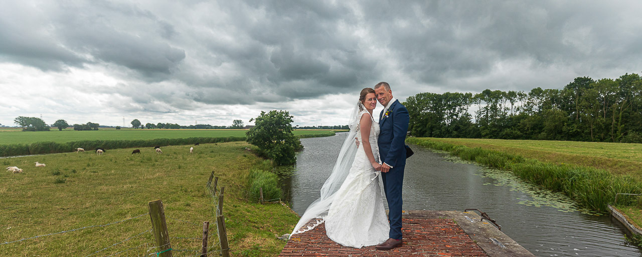 Bruidsfotografie; trouwfotograaf; Een liefdevolle pose van het bruidspaar met op de achtergrond het Middag-Humsterland en het Aduarderdiep, onder een mooi bewolkte hemel