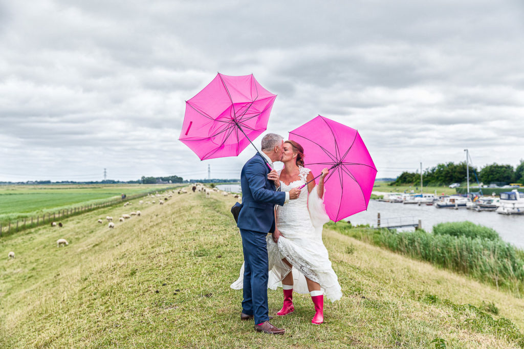 Bruidsfotografie; trouwfotograaf; Bruidspaart poseert met roze paraplues en roze regenlaarzen op de dijk langs het Reitdiep, in Garnwerd.